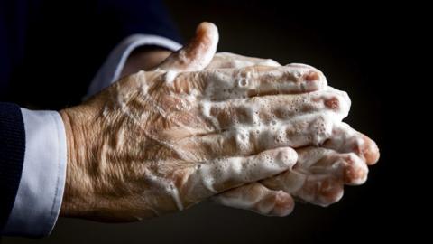 A close up image of the hands of a person who is wearing a suit. The picture has been taken while they are washing their hands and there is soap residue which is yet to be washed off.