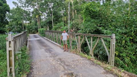 A man stood on a road bridge in what looks like a forest. He is wearing a grey t-shirt and green shorts with flip flops on.  
