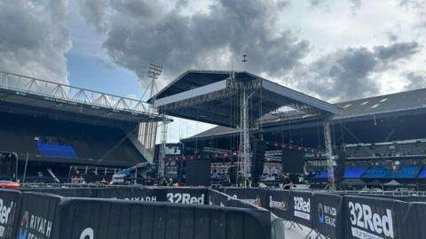 Large covered stage in the centre of the image. There are two football stands in the background. The sky is blue with moody grey clouds coming in above the stadium. 