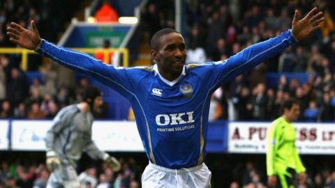 jermain defoe holds his arms outstretched wide with a small smile on his face. he wears a long sleeve blue shirt with gold trim and white collar.