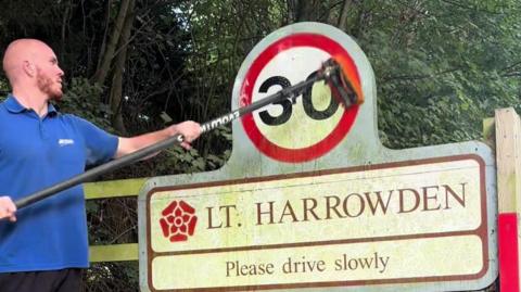 A man in a blue polo shirt cleaning a "30" sign. The 30 is black on a white backgound with a red circle around it. It is above another sign saying "Lt Harrowden" on one line and "please drive slowly" on the next. He is cleaning it with the long-handled brush. Behind the sign is trees.