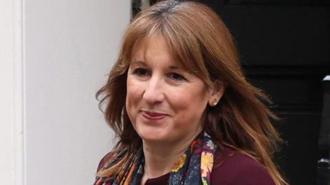 A smiling woman with shoulder-length brown hair is wearing a plum coloured jacket and floral scarf