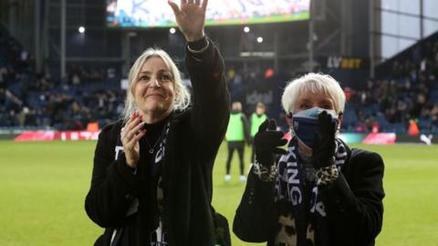 Dawn Astle with her mother and widow Larraine Astle on the pitch at the Hawthornes Stadium before West Bromwich Albion's game against Peterborough United. She and her mother are wearing black coats and are waving to the crowd