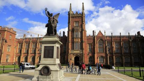 The exterior of Queen's University Belfast, a large gothic redbrick building with a statue in the font of an angel. There are some people 