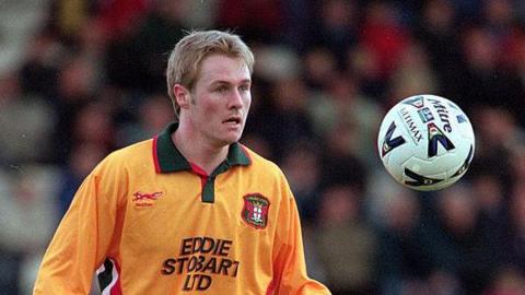 Tony Hopper on the pitch wearing an orangey-yellow Carlisle United away top as worn between 1998 and 2000. He has short blond hair and is staring towards a ball in the air at about shoulder height. The crowd is blurred in the background.