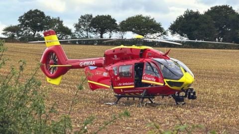 A red helicopter with yellow markings on the ground in a grassy field with trees in the background