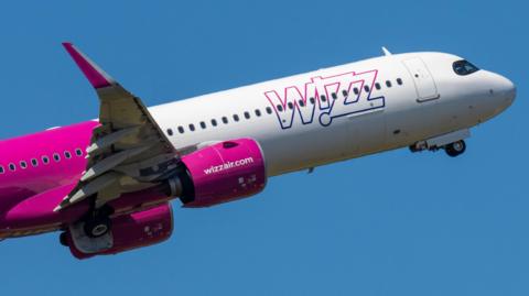A Wizz Air plane is climbing into a blue, sunny sky after take off with its wheels being retracted.