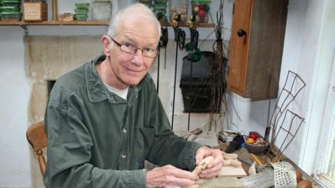 Robert Race sitting at a desk. He has short white hair, black-rimmed glasses and is wearing a dark-green corduroy shirt. He is smiling at the camera while holding some pieces of a wooden toy in his hands. 