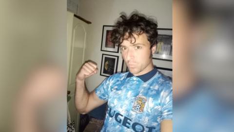 A man with brown hair wearing a sky blue Coventry City shirt form the early 1990s