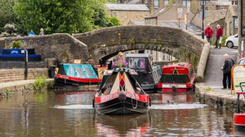The Leeds Liverpool Canal in Skipton, Norh Yorkshire, with a stone bridge over the canal in the distance and a number of colourful barges on the water