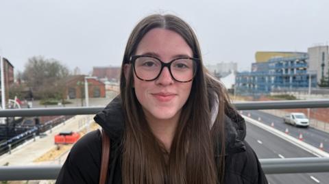 A bespectacled woman standing on a bridge over a busy road smiling. She has long dark hair and is wearing a dark-coloured jacket.