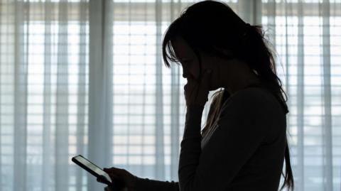 A silhouette of a woman holding her hand to her mouth and the other hand is holding a phone. She is staring at the phone. 