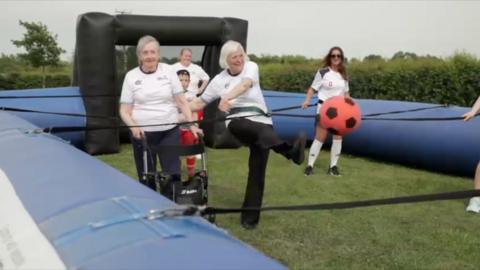 Two female care home residents taking part in a game called human table football they are inside an inflatable games course and are attached to one another with bungee cords. There are people in the background and one of the women is kicking a red football.