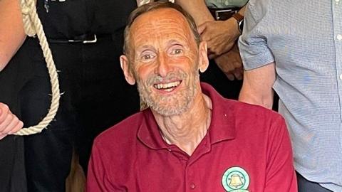 Mike Wigney looks and smiles at the camera. He is an older man with short brown hair and a beard.