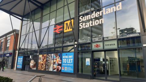 Sunderland Station is a tall glass building with sliding doors. A large sign above the entrance reads Welcome to Sunderland Station. The Tyne and Wear Metro's yellow logo is next to it. Advertising posters cover the bottom windows showing photos of coffee and food and reading Retail Unit To Let. 