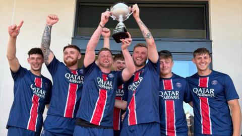 Seven men in a dark blue football kit with a red vertical stripe down the middle of it are celebrating. The two men in the centre are holding up a silver cup. They are al smiling and some of them have their hands in the air.