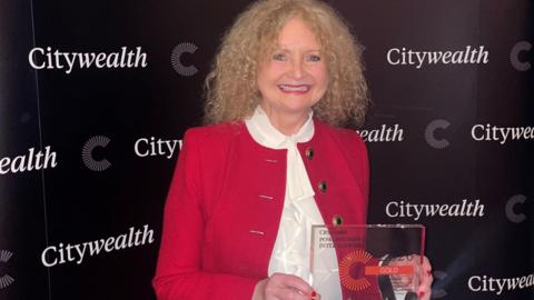 A woman called Kay McCarthy holds an award she has won at a ceremony in London. She has a broad smile on her face and is wearing a red blazer with a white blouse underneath. McCarthy has long tight blonde curly hair. A sign behind her says "Citywealth". The award is mainly made from glass.