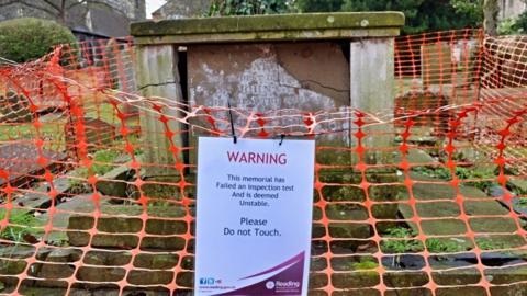 Warning sign attached to orange net-style fencing with stone tomb in churchyard behind