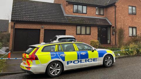 A police car is outside a red brick house with a double garage in Lingwood Park in Peterborough. Around the house is blue and white tape as a police cordon. 