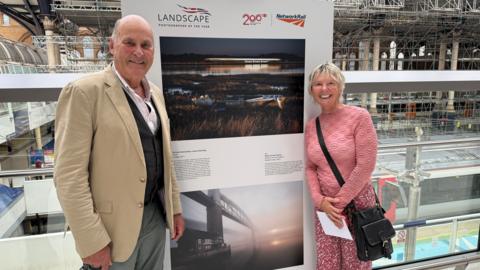 Charlie Waite, wearing a shirt, waistcoat and jacket stands in front of a photo on display. Also next to the photo is Elizabeth Coles wearing a pink jumper and pink patterned trousers.