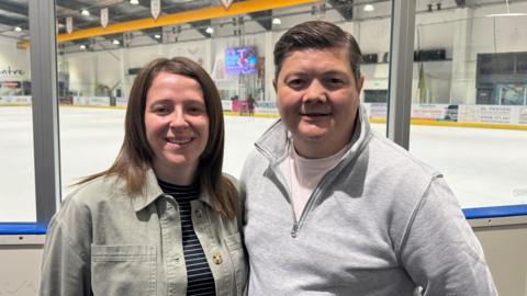 Joanne and Jason Parry (left to right). They are standing close together, with an ice rink in the background. Joanne has dark brown shoulder length hair and is wearing a khaki shirt over a striped black and white top. Jason is wearing a grey jumper with a white t shirt seen underneath. He has light brown hair.