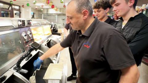 Behind the counter at Ruby's Chippy in Thringstone, two young men watch as an older man reaches to drop something in to big deep fryer.