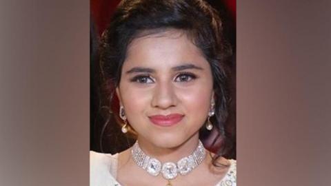 A smiling woman with tied back black hair wearing gold earrings and a diamond looking necklace and white dress 