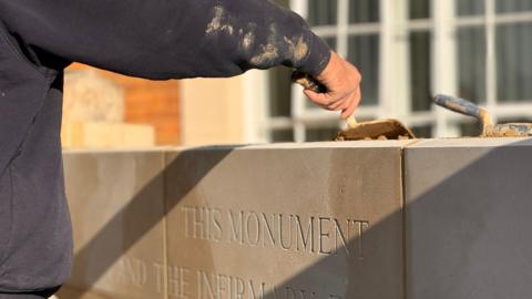 The hands of a person can be seen laying cement on top of a white stone structure. An inscription can be partially seen, reading: "This monument".