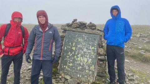 Mae tri dyn ifanc mewn siacedi gaeaf coch, llwyd a glas yn sefyll ar gopa mynydd wedi'i orchuddio â chymylau
