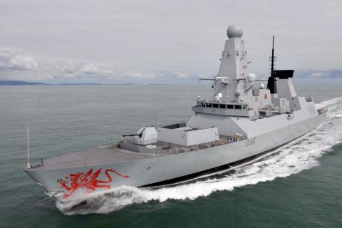 HMS Dragon at sea.  The warship is a Type 45 Destroyer, grey in colour with a red dragon on the bow. She is pictured at sea against a backdrop of grey and cloudy skies