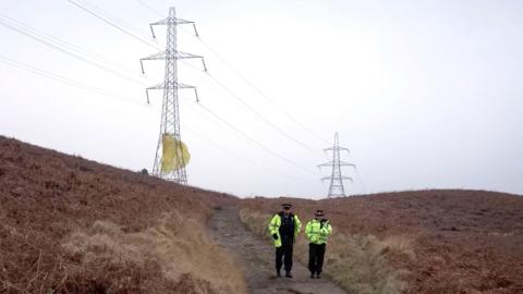 A male and a female police officer walk down a remote dirt track, which is flanked on both side by gorse. On the horizon are two large electricity pylons, there is yellow-coloured material that is wrapped around the bottom of the nearest pylon that appears to be billowing in the wind  