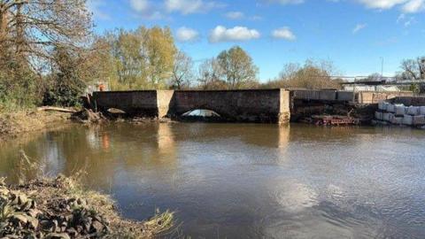 A low brick bridge over a river is pictured with trees at one end and scaffolding and sand bags at the other where it has been damaged. The underside of the bridge is a series of arches. 