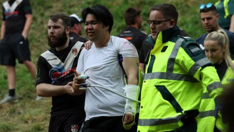 A man with an arm wrapped in bandages is helped away from Cooper's Hill by a paramedic and another competitor following a fall at the Gloucestershire cheese rolling event.