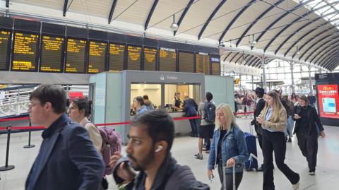 Passengers walk past the ticket and information kiosk. It is a grey booth with a clear screen and is positioned in front of several electronic departure boards.