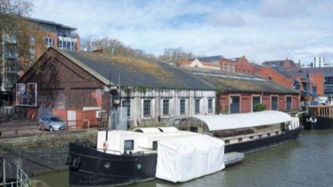 A long barge with a black bottom and white rounded shelter over the top. It is floating in the harbour beside an industrial brick shed. The barge looks in a state of some disrepair.