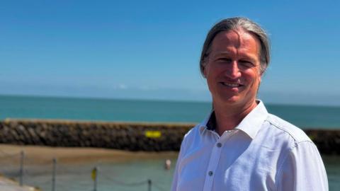 A smiling man with tied-back long grey hair wearing a white shirt. He is standing next to the sea.