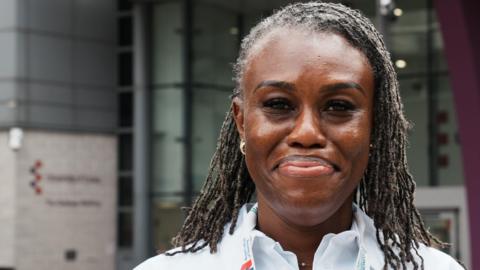Ijeoma Okolo wears a white shirt with a lanyard around her neck. She has braided hair and is smiling at the camera. She stands in front of the Gateway building at the University of Essex's Southend campus.
