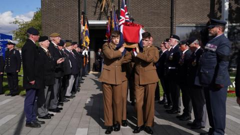 Young men in uniform carry a union jack draped coffin out of a church past veterans, also wearing uniform, during a funeral.