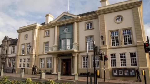 Ulverston's Coronation Hall - a three-story Edwardian theatre, with five windows on either side of the ground floor entrance, three on either side on the first floor, and two on the second. It is pale coloured with darker stone facing, and green on the section above the entrance door, which has a column on either side. To the right is a statue of Laurel and Hardy.