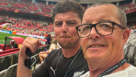 Two football fans in a stadium. The one nearest the camera has dark/grey receding short hair and glasses. The one behind his is younger nad has dark spiky hair and it in a black New Zealand top and is holding sunglasses in his right hand.