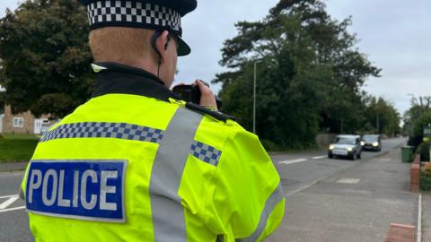 A police officers wears a high vis vest and stands on the pavement taking pictures of speeding cars