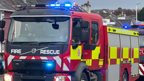 A moving red and yellow fire engine with blue lights on top of it. On the front of it has the words 'fire rescue'. Inside the vehicle there are two people in vision with on driving the engine. 