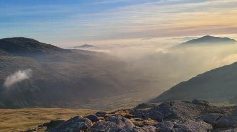 The Duddon Valley which is surrounded by fells. There is a layer of fog covering the valley and obscuring the peaks in the distance. The sky is blue on the left and an orange on the right with wispy clouds.