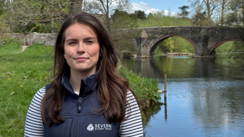 A woman with long dark hair and a blue top with blue and white striped sleeves standing in front of a river with a stone bridge with a number of arches going over it