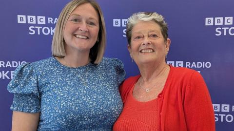 Two women standing together and smiling. The woman on the left has blonde bob hair style and the woman on the right is wearing a red top and red cardigan. She has short hair which is black with a grey fringe. 