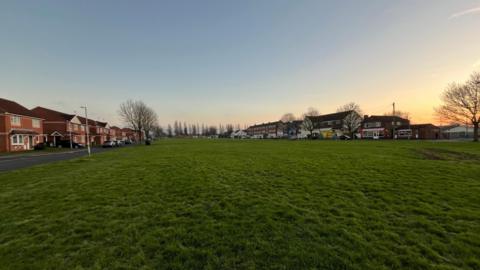 A large green space in an urban area during a clear winter sunset. There are houses and shops surrounding the green space, which is mostly just an empty field, with trees in the background