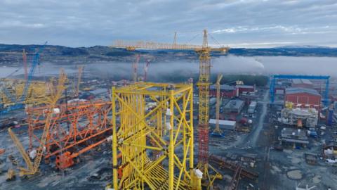 Drone footage of an industrial oil and gas construction site, with enormous yellow and orange leg sections and cranes and sheds in the background. 