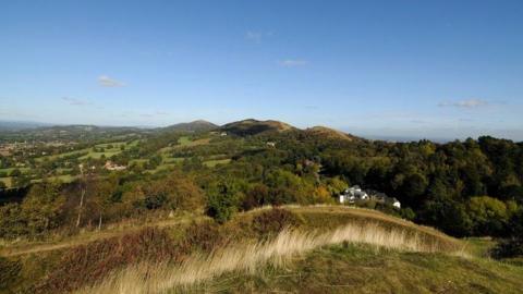 A view from on top of the Malvern Hills on the border of Worcestershire and Herefordshire. There are white buildings in the distance, with hilltops further away, and there is woodland around the houses.
