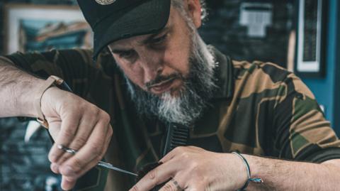 Dan James cutting hair at his Minehead salon. He has a beard and is wearing a cap