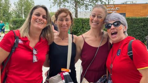 Four women looking into the camera, smiling. Ones on the left and right of the group are wearing red t-shirts, either side of a woman wearing a black vest top and one with a burgundy vest top. They all have small Spanish flags in face paint on their cheeks.